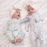 Two babies wearing floral outfits and headbands lying on a textured white surface.