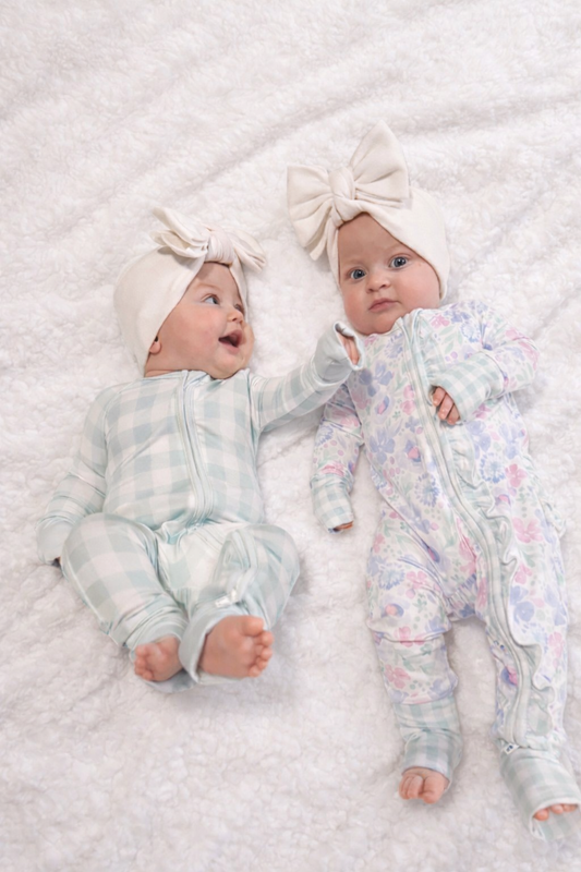 Two babies wearing floral outfits and headbands lying on a textured white surface.